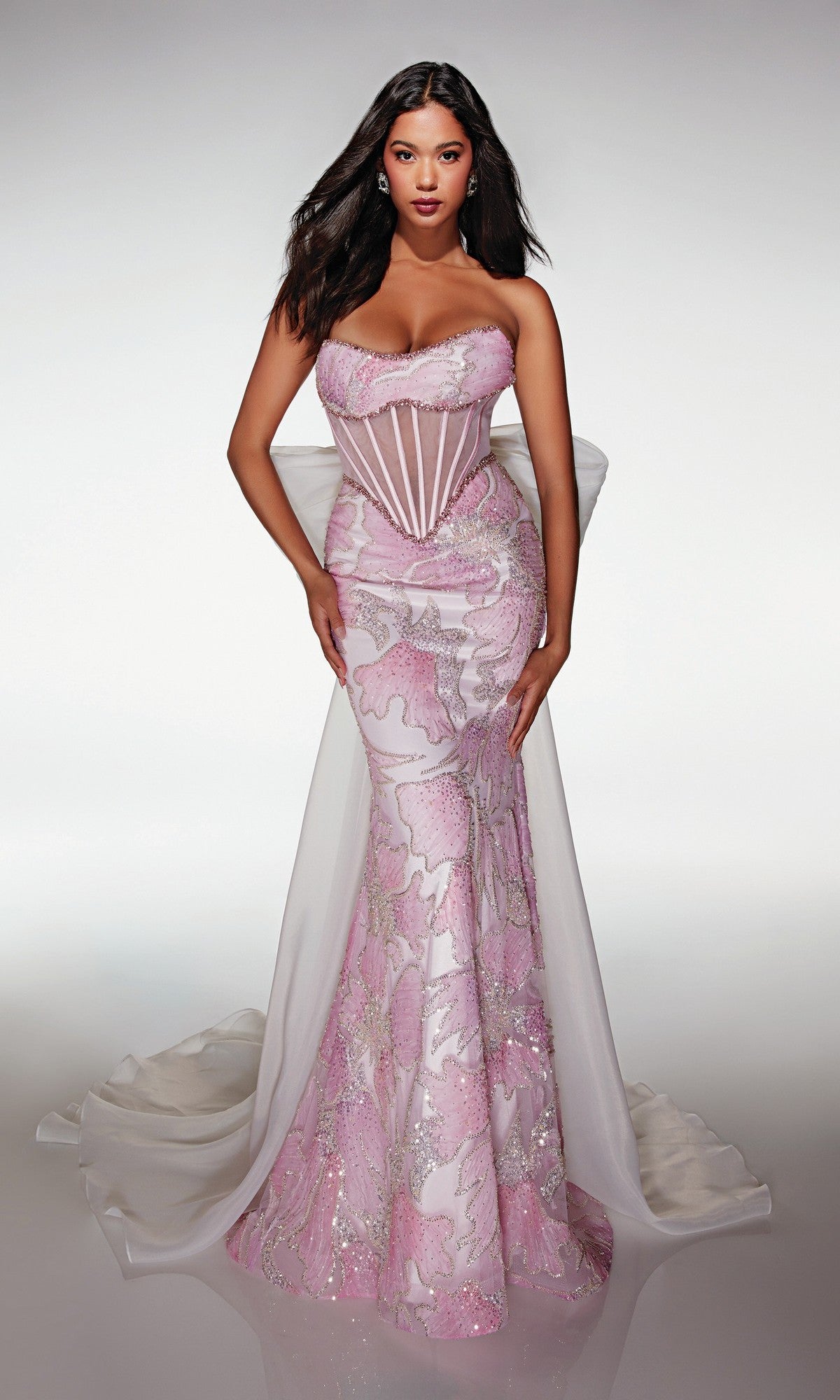 A woman wearing the Alyce 62187 long prom dress in pink/white, featuring a strapless, form-fitting silhouette with a sheer corset bodice, lace accents, and a long white train, stands against a plain light background.